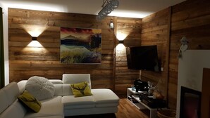 En hyggelig stue med en hvid sofa, to dekorative puder, et ophængt TV og et maleri af et bjerglandskab.