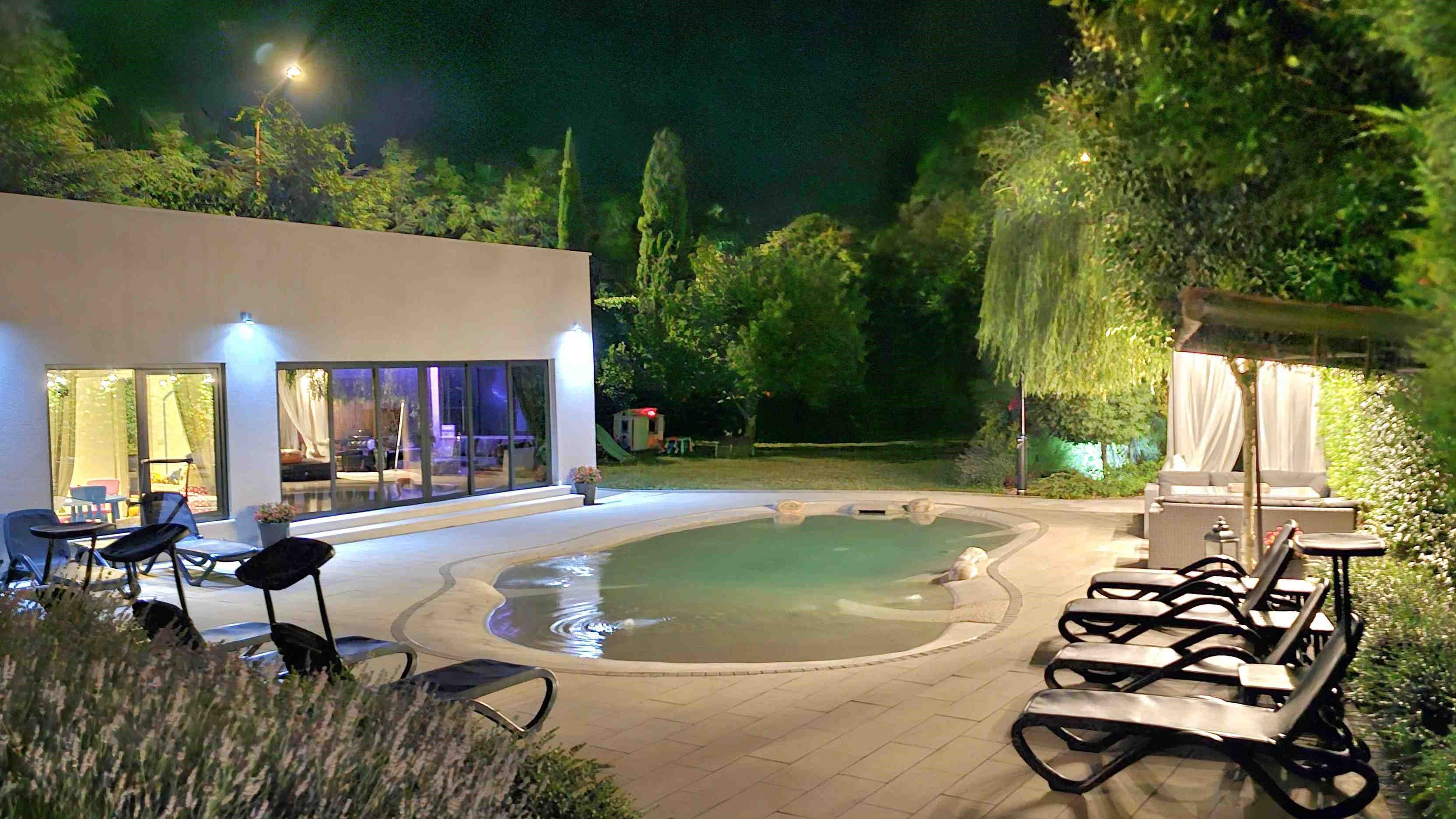 Un área de piscina exterior de noche con sillones reclinables, un edificio moderno con puertas de vidrio y abundante vegetación circundante.