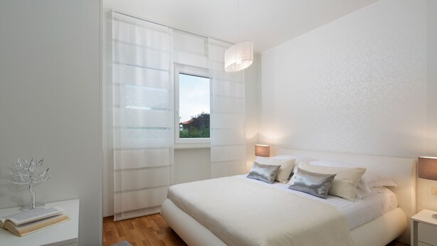 Un dormitorio moderno con una cama grande, lámparas de noche, una ventana con cortinas traslúcidas y una mesita de noche con un libro y un objeto decorativo.