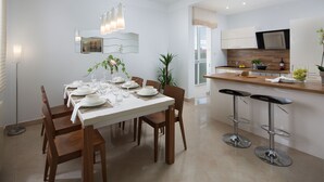 Una cocina moderna con armarios blancos, encimera de madera y horno empotrado. Hay un comedor con una mesa blanca dispuesta para seis comensales, sillas de madera y una lámpara colgante.
