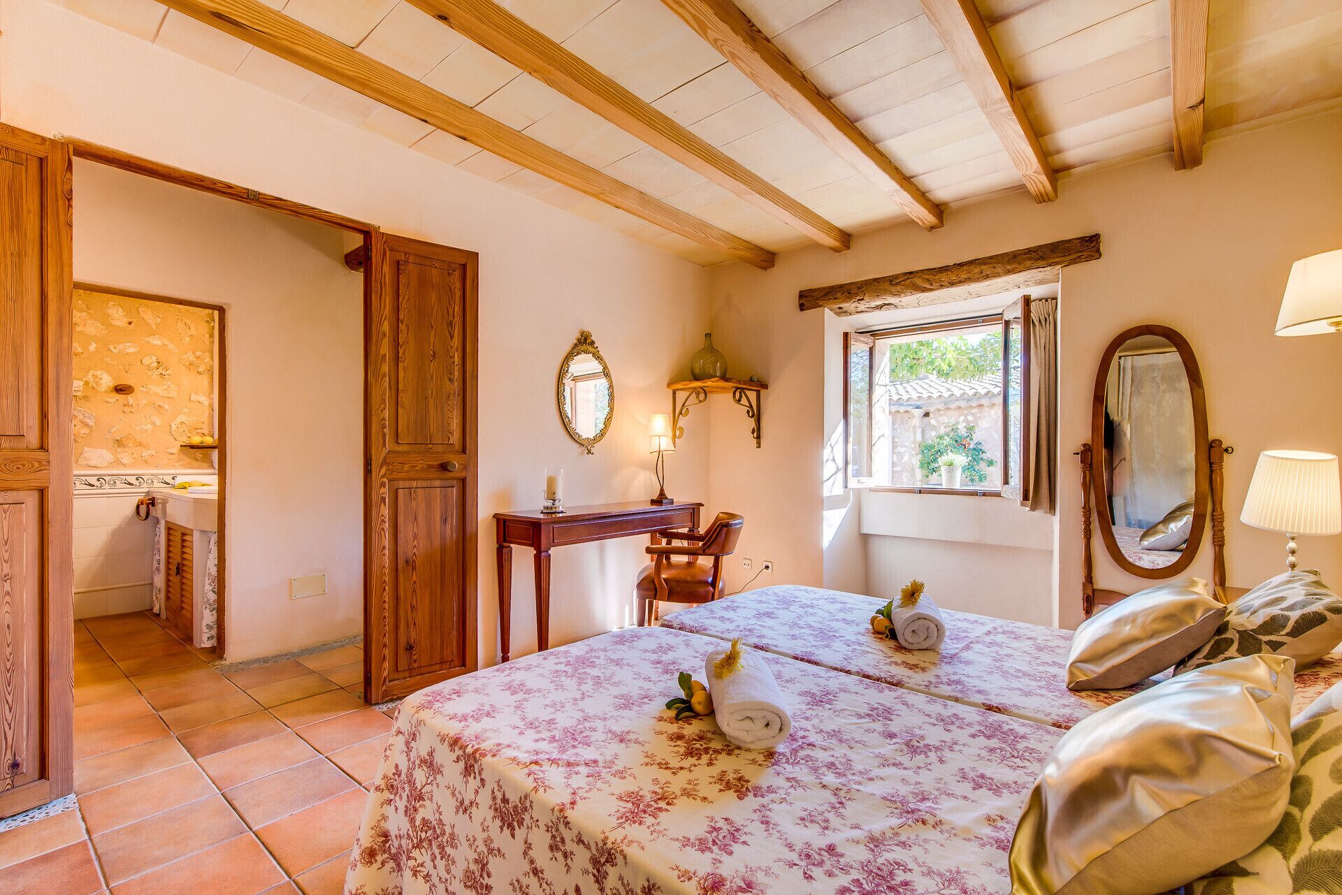 Une chambre avec un plafond en bois, un lit recouvert d’une literie à motifs floraux, une table de chevet avec une lampe et une fenêtre donnant sur un paysage verdoyant.