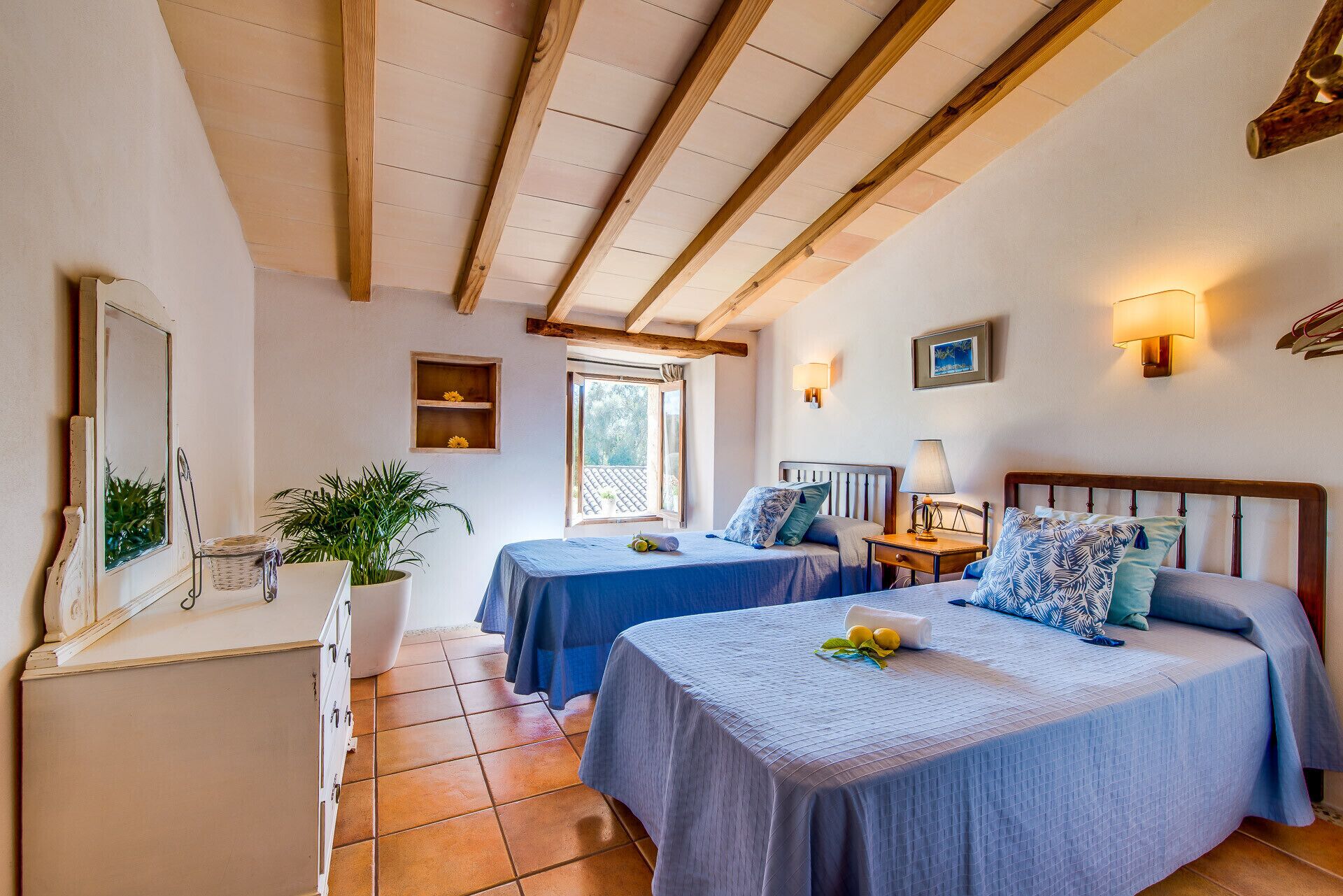A room with two beds, a wooden ceiling, a window, a plant, and a wall-mounted lamp.