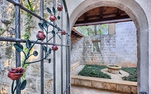 A courtyard with a stone wall, a metal gate with red flowers, and a fountain.