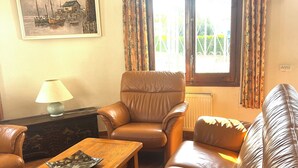 A living room with a brown leather sofa, a wooden coffee table, and a leather armchair. There is a painting on the wall and a window with curtains.