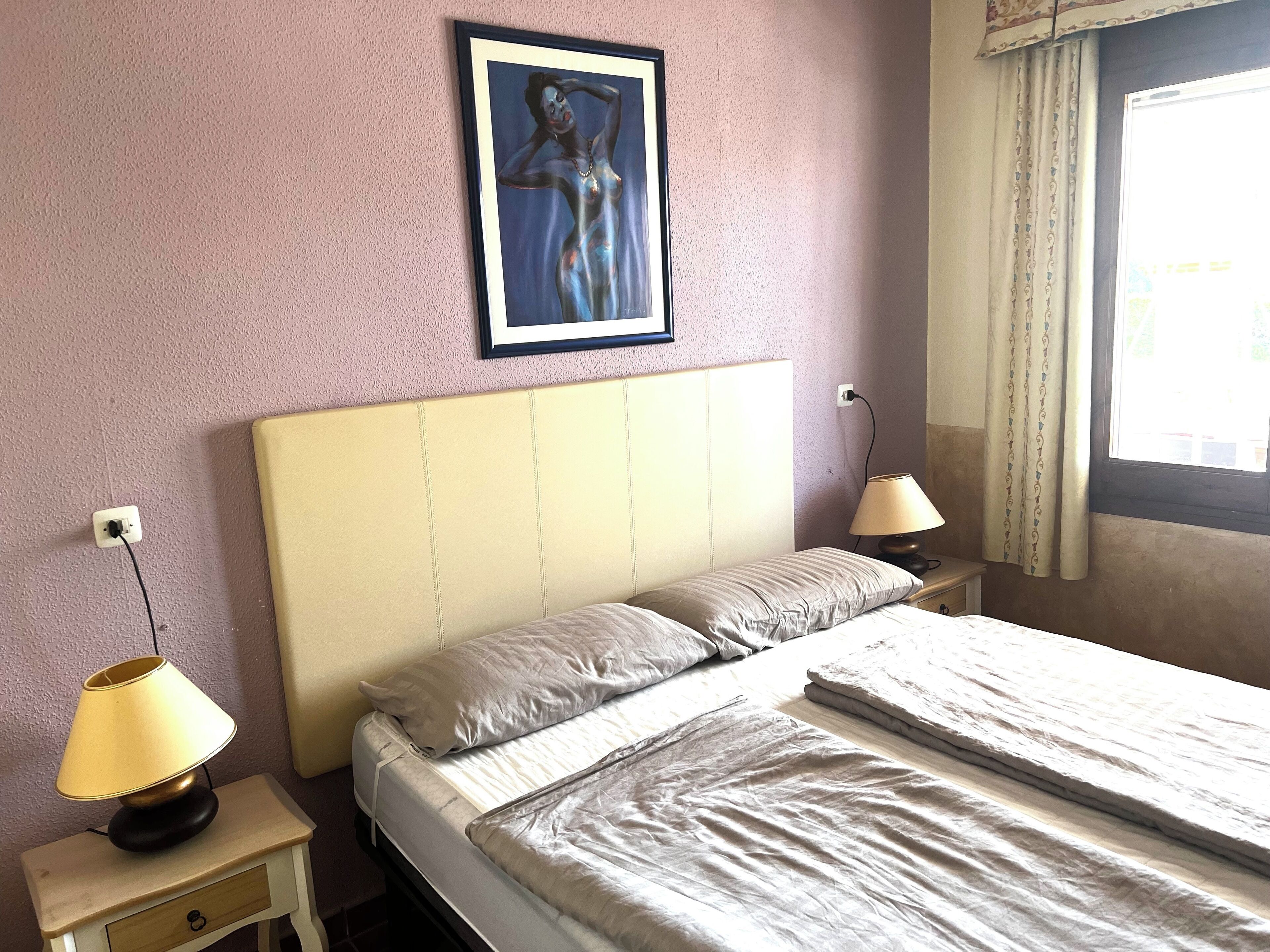 Ein Schlafzimmer mit einem Bett, zwei Nachttischlampen, einem Nachttisch, einem gerahmten Bild an der Wand und einem Fenster mit Vorhängen.