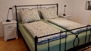Ein Schlafzimmer mit Metallbettgestell, grüner Bettwäsche, zwei Nachttischen mit Lampen, Holzboden, einem an der Wand hängenden Bild und einer Pendelleuchte.