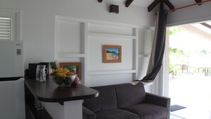 A modern kitchen with a dark countertop, a refrigerator, and a bowl of fruit. There is a gray couch and a framed picture on the wall.