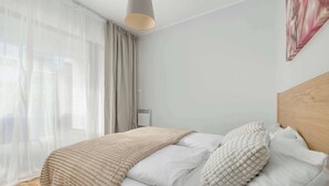 A neatly made bed with a beige blanket and white pillows, a wooden headboard, and a large window with sheer curtains.