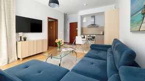 A modern living room with a blue sofa, a glass coffee table, a TV, and a kitchen in the background.