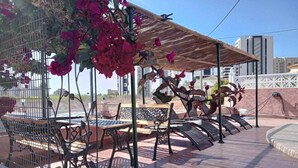Uma área externa com assentos de metal, cadeiras e uma mesa de madeira, cercada por uma grade de metal e uma pérgola com plantas trepadeiras.
