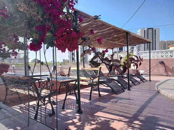 An outdoor seating area with metal chairs and a wooden table, surrounded by a metal fence and a pergola with climbing plants.