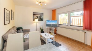 Una sala de estar moderna con sofá, mesa de comedor, televisor y vista al exterior a través de amplias ventanas.