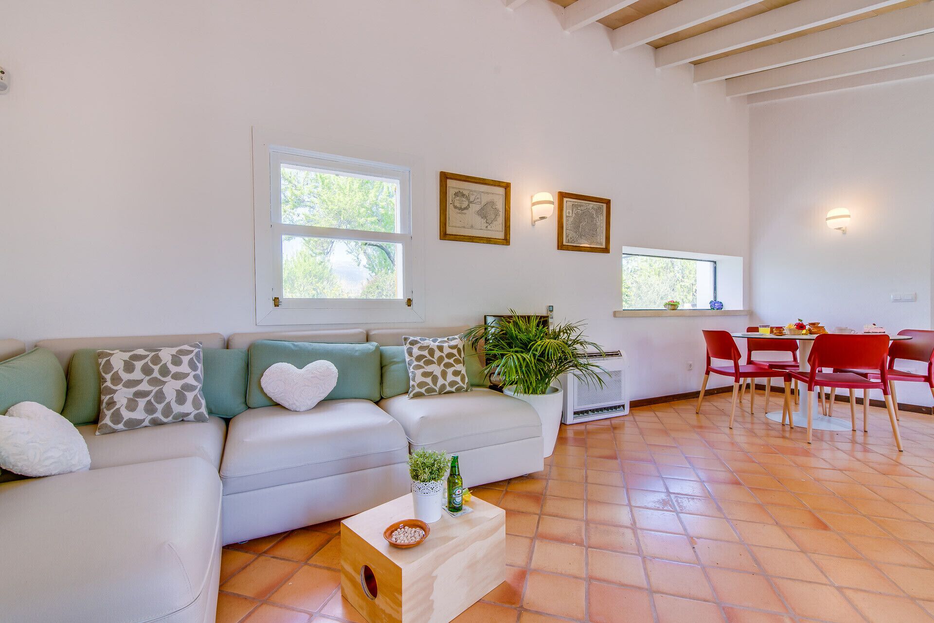 Uma sala de estar moderna com um sofá branco, cadeiras vermelhas e uma mesa de jantar. Há duas janelas com obras de arte acima e uma planta em vaso no chão.