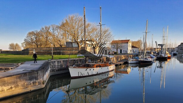 A calm harbor with several boats docked, a person walking on a path, and a building with a red roof in the background.