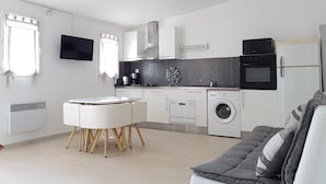 A modern kitchen with white cabinetry, a built-in oven, a microwave, and a washing machine. There is a dining table with chairs and a sofa in the living area.