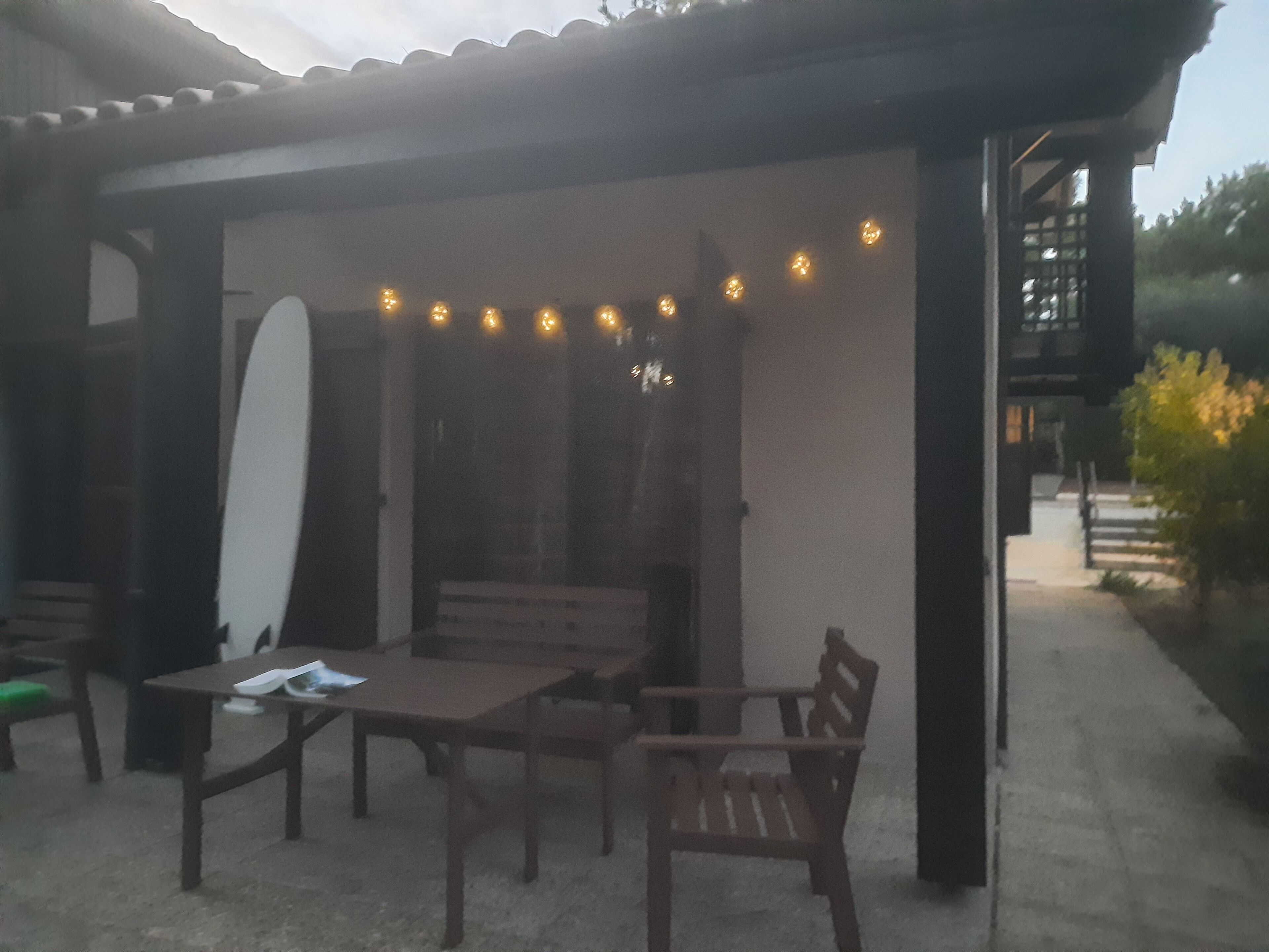 An outdoor seating area with a table and chairs, a surfboard leaning against a wall, and string lights above.