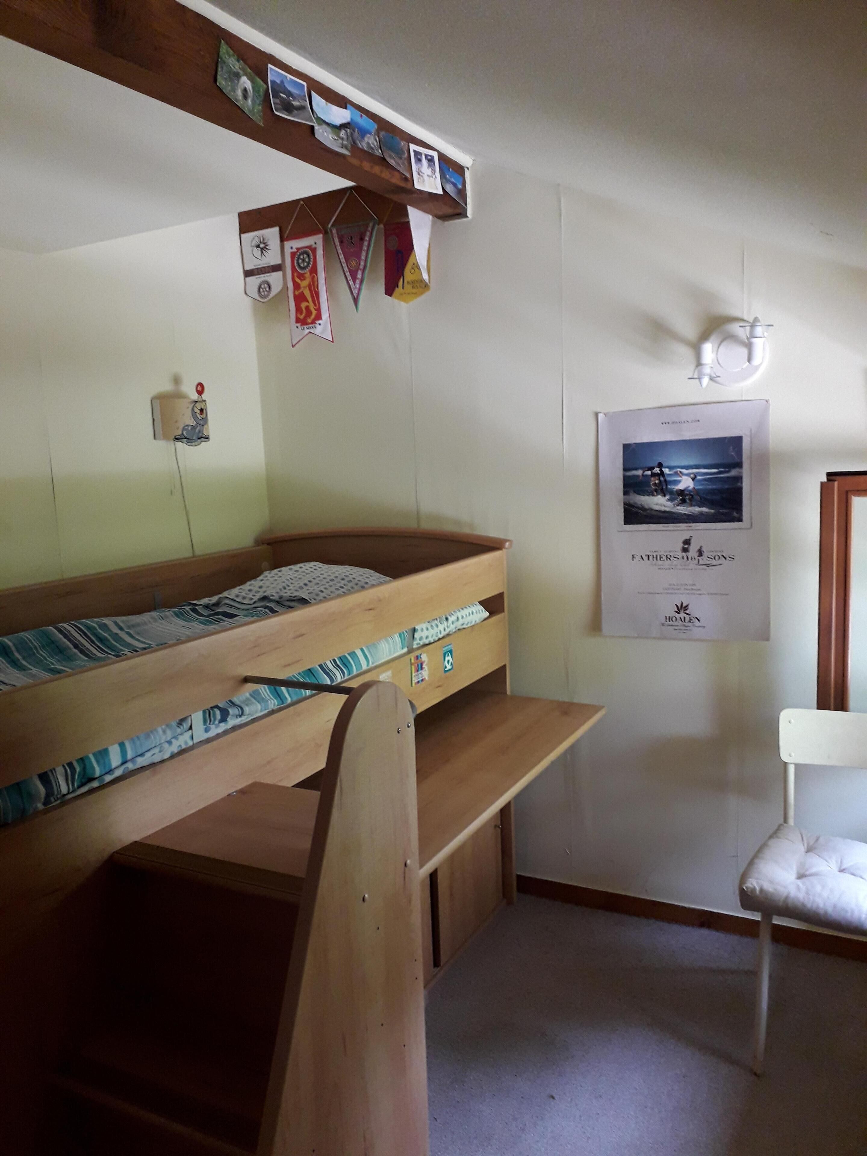 A bunk bed room with a wooden headboard, a small desk, and a chair. There is a poster on the wall and a shelf with photos.