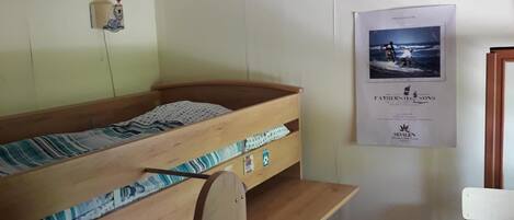 A bunk bed room with a wooden headboard, a small desk, and a chair. There is a poster on the wall and a shelf with photos.