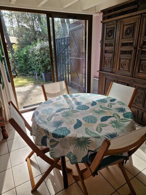 A dining area with a round table covered by a tropical leaf-patterned tablecloth, surrounded by wooden chairs. A sliding glass door leads to a garden with a wooden fence and trees.