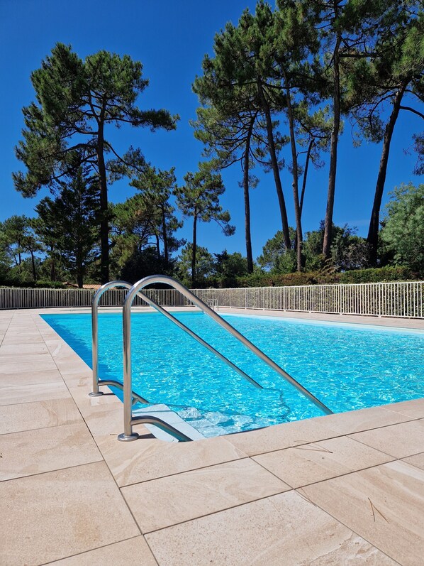 A swimming pool with a metal ladder, surrounded by a tiled deck and tall pine trees. - House with shared pool 30m far, 100 m from the Golf de l'Ardilouse (Lacanau Océan)