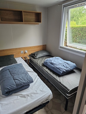 A compact room with two beds, a wooden shelf, a window with a view of greenery, and a power outlet.
