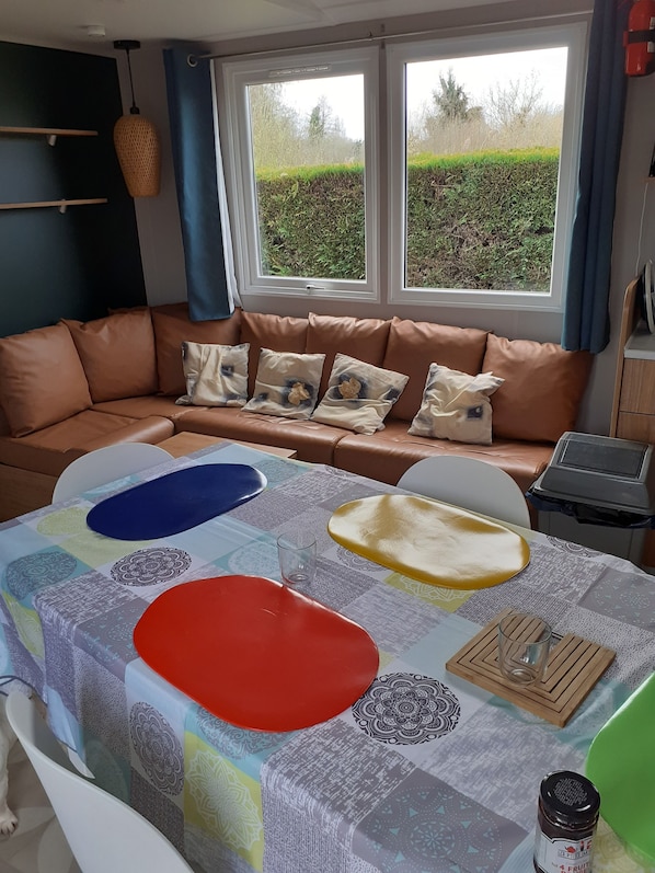 Un salon chaleureux avec un canapé en cuir marron, une nappe colorée et une vue sur la verdure à l’extérieur.