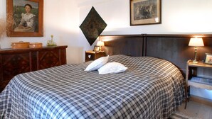 Una cama bien hecha con una colcha a cuadros, dos almohadas y un cabecero. Hay dos mesitas de noche con lámparas, un cuadro enmarcado en la pared, y un aparador de madera con un jarrón y una tetera encima.