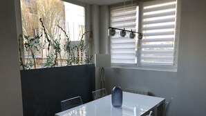 Zona pranzo con tavolo e sedie bianche, una lampada a sospensione e vista attraverso una finestra con persiane.