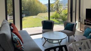 Un salon moderne comprenant un canapé, deux fauteuils, une table basse et un téléviseur. De grandes portes-fenêtres coulissantes donnent sur un jardin avec une pelouse et des arbres.