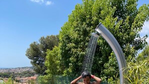 Une personne se tient sous une pomme de douche sur une terrasse en bois, avec une piscine en arrière-plan.