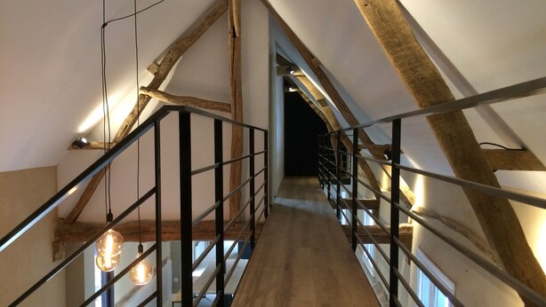 Een houten trap met metalen leuningen en zichtbare houten balken, leidend naar een donker interieur.