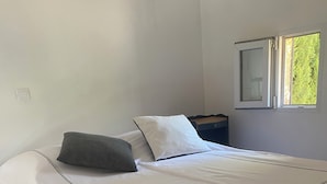 Una cama bien hecha con cubrecama y almohadas blancas, suelo de madera y ventana con vistas a zonas verdes.