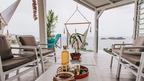 Un espace extérieur aménagé avec une zone de repos couverte, comprenant des chaises blanches et une table, un hamac et une vue sur la mer.
