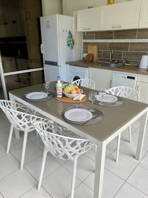 A dining table set with plates, cutlery, and a bowl of fruit, surrounded by white chairs in a modern kitchen with white cabinets and a dishwasher.