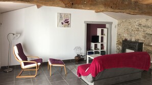 A cozy living room with a wooden ceiling beam, a sofa, a chair, a small table, and a television on a white shelf. - DOMAINE DE LA MORINIERE - THE STABLE (Commequiers)