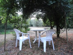Una mesa blanca de plástico y dos sillas bajo un dosel de árboles en un entorno natural.