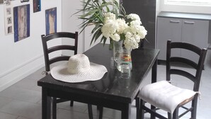 Comedor con una mesa negra, sillas blancas y un florero con flores blancas. Hay cuadros en la pared y una ventana con persianas.