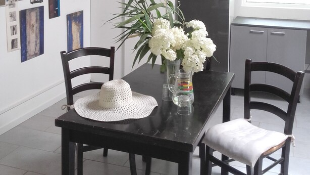 Un coin repas avec une table noire, des chaises blanches et un vase rempli de fleurs blanches. Des tableaux encadrés ornent le mur, et une fenêtre est équipée de stores.