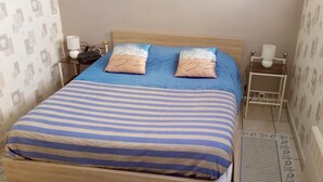 Una cama bien hecha con una colcha azul y blanca a rayas, dos almohadas con diseños de corazón, y un cabecero de madera. Hay dos mesitas de noche con lámparas y un pequeño estante.