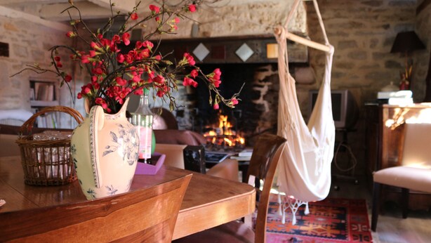 Un interior rústico con una mesa de madera, una silla colgante y una chimenea al fondo.