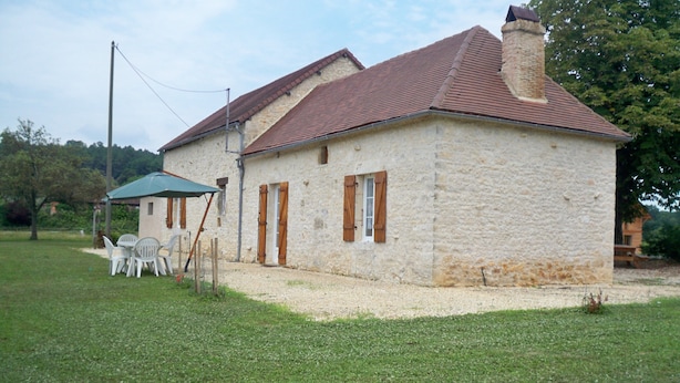 A stone building with a red-tiled roof, a small porch with chairs, and a gravel driveway.