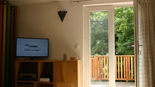 Un salon avec un téléviseur posé sur un meuble en bois, une porte-fenêtre coulissante donnant sur une terrasse en bois et un élément décoratif fixé au mur.