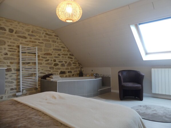 A bedroom with a stone wall, a sloped ceiling, a skylight, a bed, a chair, and a built-in bathtub. - Luxury cottage  (SAINT EVARZEC)
