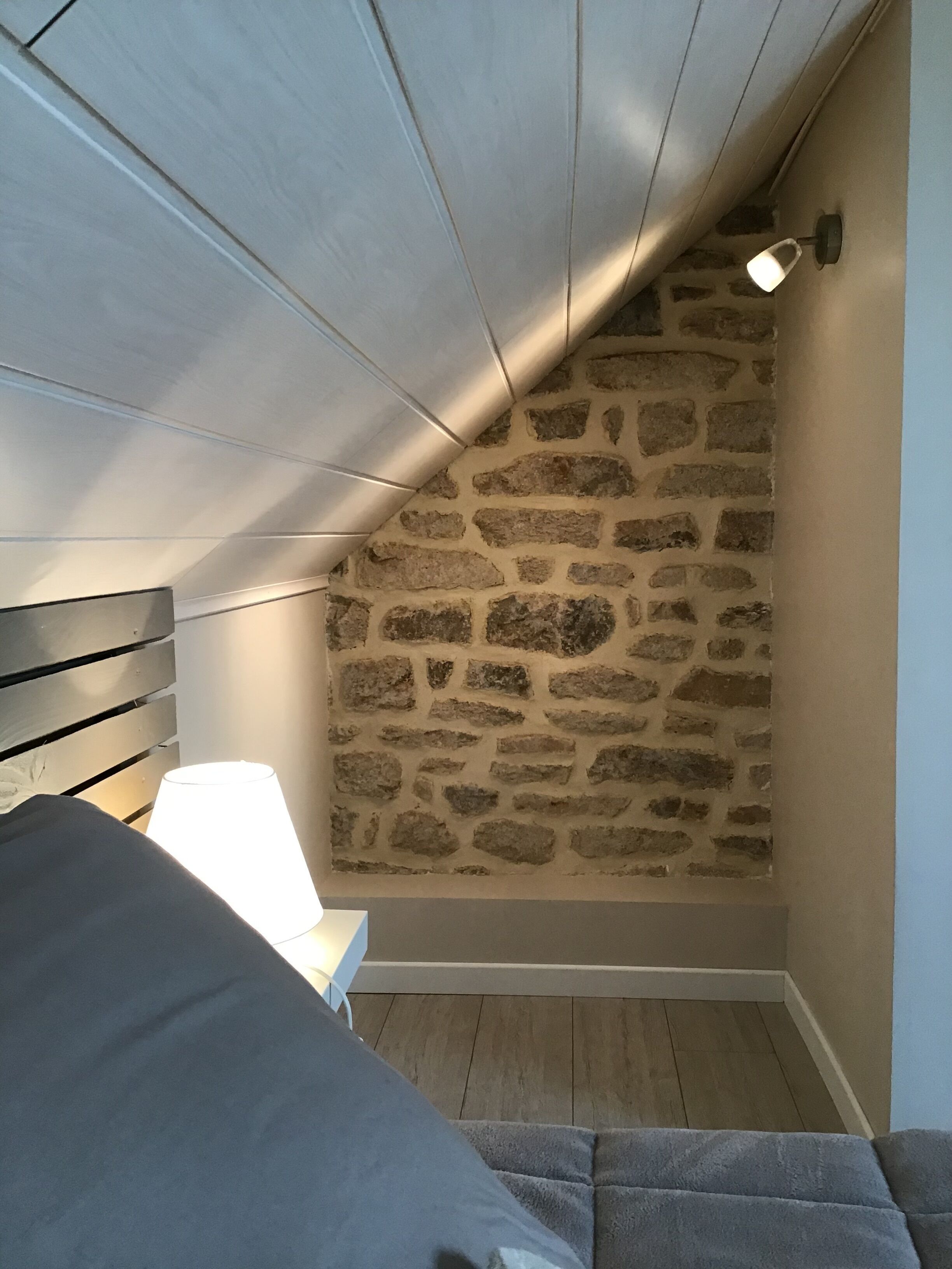 A bedroom with a stone wall, a wooden sloped ceiling, a bed, and a nightstand with a lamp.