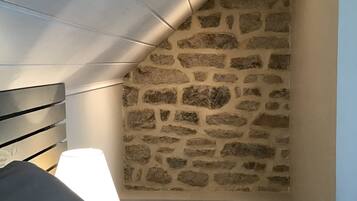 A bedroom with a stone wall, a wooden sloped ceiling, a bed, and a nightstand with a lamp.