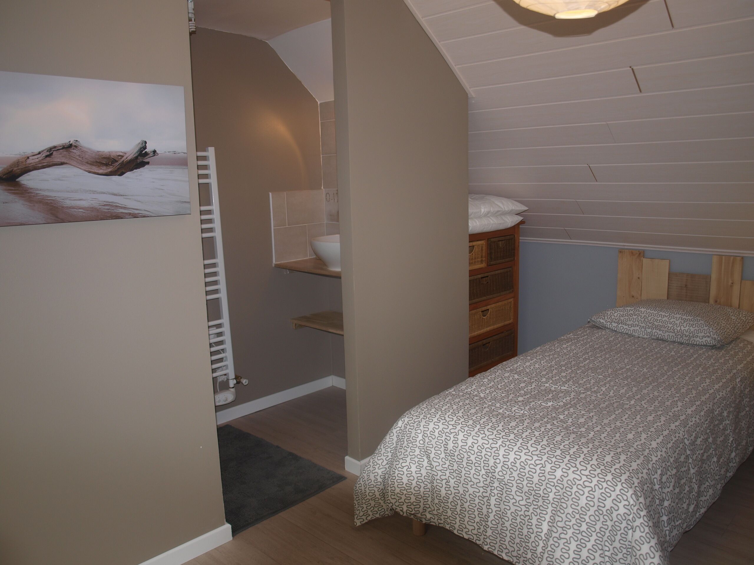 A bedroom with a sloped ceiling, a wooden headboard, a bed with patterned bedding, a white wall-mounted shelf, and a painting of a tree branch.