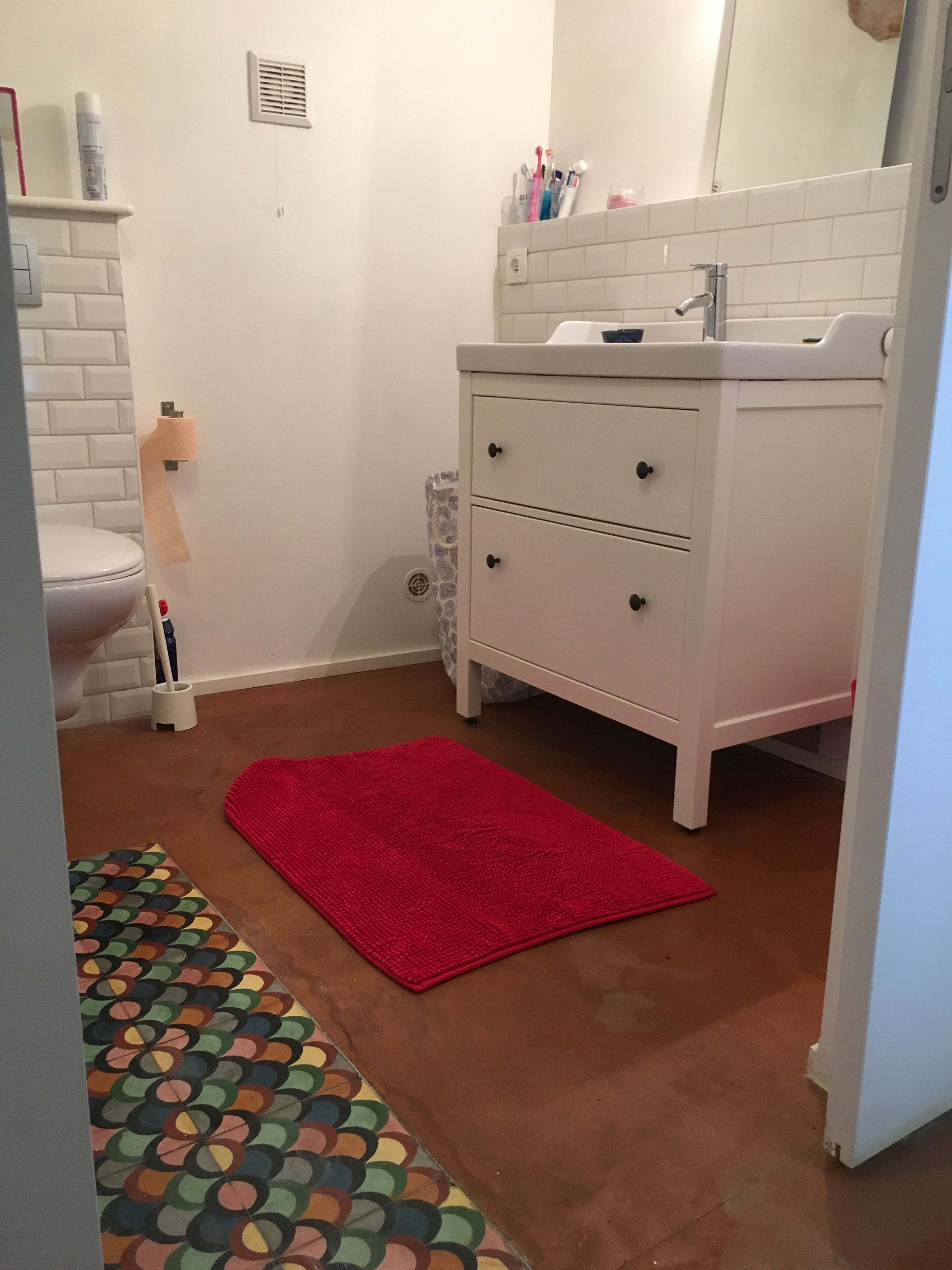 A bathroom with a toilet, a white sink with a mirror, a red rug, and a patterned rug on the floor.