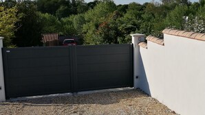 Ein verschlossenes Tor mit schwarzem Gitter, eine weiße Mauer mit einem Ziegeldach und ein Schild.