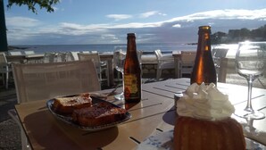 Ein Tisch mit Essen, zwei Flaschen, einem Glas und einer Nachspeise, vor dem Meer an einem sonnigen Tag.