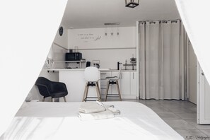 A modern, minimalist room with a bed, two chairs, a small kitchenette, and a curtain-covered window. - Gîte *** 🌿 Le Mesminois - Well-being, nature and comfort just a stone's throw from Puy du Fou (Saint-Mesmin)